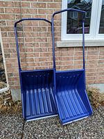 Front view of two large blue snow shovels standing side by side against a brick wall, showing metal handles and wide plastic scoops.