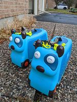Two blue dog-shaped ride-on suitcases side by side, showing front faces with eyes, ears, nose, and bone-shaped locks on top