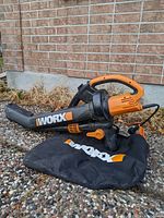 Full view of WORX electric leaf blower with attached black collection bag on gravel surface alongside brick wall.