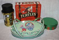 View of four collectible tins on white cloth background, showing various designs including Nestle Turtles, Mr. Peanut, a small striped round tin, and an Easter tin