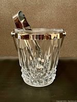 Side view of the gold-rimmed crystal ice bucket showing the detailed cut crystal design and metal rim with attached handle, along with the metal ice tongs placed inside.