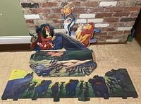 Photo of three cardboard Lion King advertising pieces: a character cutout and two scenic panels with animal silhouettes, laid out on a tiled floor against a brick wall.