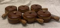 Set of 6 brown ceramic French onion soup bowls with matching lids and handles, arranged on a cloth surface.