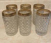 Set of six clear crystal highball glasses with diamond-cut pattern and gold decorative bands on rims on a marble surface.