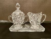Glass creamer, sugar bowl with lid, and rectangular tray on a dark background, showing detailed pinwheel and starburst cut glass patterns.