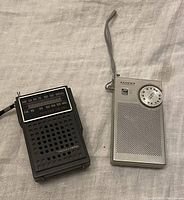 Photo of two transistor radios on a light fabric background, one black Realistic and one white Lloyds