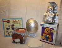 Four cake pans together: Betty Crocker Bake Art springform pan set in box, Winnie the Pooh aluminum cake pan standing on box, metallic Easter egg pan, and a white-colored decorative item shaped like a heart in front.