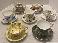 Overview of the seven tea cups and seven saucers with various floral and leaf designs from different brands.