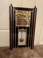 Full view of large rectangular wall clock with black and gold striped mirrored frame, square clock face with roman numerals, and visible pendulum underneath.