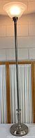 Full view of tall standing chrome and steel torchiere floor lamp with frosted glass shade on top.