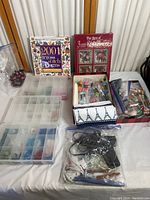 Full lot overview showing two cross stitch books, multiple plastic organizer boxes with beads, crafting tools, and small decorative items on a white surface.