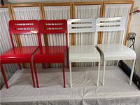 Four metal stacking chairs, two red and two white, arranged side by side against a backdrop with curtains. Chairs have horizontal slots on backrests and solid seats, with straight legs and glossy finish.