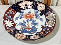 Top view of large Japanese Imari porcelain plate showing central floral motif, surrounding detailed flowers in red, blue, white and gold with gold rim.