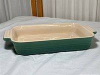 Side angled view of Le Creuset green and cream rectangular casserole dish placed on white cloth surface showing handles and detailing.