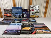 Eleven hardcover books arranged on a flat surface, showing a variety of Porsche and Mercedes-Benz covers featuring different models.