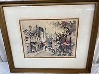 Framed print showing overall artwork with softly colored Paris street scene, cream mat, and gold frame.