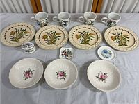 Full view of collection showing 4 large floral plates, 4 floral mugs, 3 small scalloped bowls with pink rose design, and two small floral trinket dishes on a grey cloth.