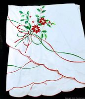 Photo showing folded white tablecloth with red scalloped edges and embroidery of poinsettia flowers and green leaves tied with red and green bows.