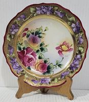 Front view of a round hand-painted ceramic plate on a wooden stand, showing floral roses and violets with gilt accents and pierced edges.