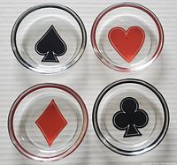 Top view of all four coasters showing playing card suits spade, heart, diamond, and club in black and red on clear glass discs.