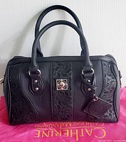 Front view of the black Catherine Malandrino handbag showing laser-cut floral patterns, handles, silver-tone clasp, and attached logo tag, placed on pink branded dustbag.
