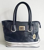 Front view of black handbag with white base trim and double handles, showing gold-tone logo plaque and round pendant charm.