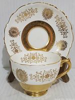 Front view of gold encrusted porcelain cup with saucer behind, showing ornate leaf and filigree gold patterning and shaped rim.