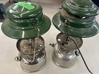 Two Coleman gas lanterns side by side showing green tops and silver fuel tanks.