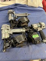 Photo showing 3 used pneumatic air coil nailers with metal bodies and black coil magazines on blue cloth background.