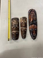 Front view of three wooden masks arranged vertically with tape measure for scale. Each mask shows distinct carved motifs including turtle, elephant, face, and fish.