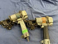 Two silver metallic Hitachi air nailers placed side by side on a blue cloth. Each has pneumatic connectors and nail feed mechanisms, showing signs of heavy use and wear.