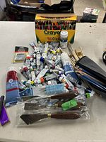 Top view of the art supplies showing oil paint tubes, brushes, Crayola crayon box, palette knives, pencils, and a bottle of paint medium or varnish on a table