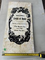 Rectangular vintage wall hanging featuring black decorative floral border and text advertising Bacorn's Cream of Roses skin-glo creme by The Bacorn Co, Elmira New York. Surface shows age-related discoloration and staining.