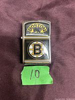 Metal lighter with Boston Bruins logos on front, placed on a cloth background with a green piece of tape marked '10' below it.