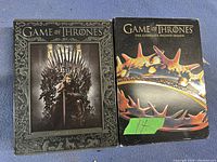 Two DVD cases for Game of Thrones seasons 1 and 2. Season 1 shows Ned Stark on the Iron Throne with a dark ornate background, Season 2 shows a golden spiked crown on black background.