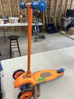 Orange children's scooter on a white table, displaying front wheels, handlebar, and platform with Little Tikes branding.