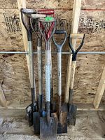Photo showing six roofing shovels with wooden handles and metal blades lined up against plywood wall