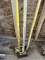 Three yellow roofing spuds leaning against a wooden beam wall showing square bases and long handles with paint wear.