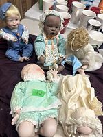 Photo shows four porcelain dolls in various conditions on a table with cups in background. Dolls include a blonde male doll in blue overalls, an African American doll in lace dress, a curly blonde doll, and a baby doll in a mint green dress.