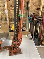 Clear image of the orange ratchet car jack standing on a white tabletop in a workshop setting, showing the height adjustment holes and lifting mechanism.