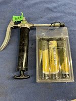 Photo of a small metal hand lever grease gun with black handle and grease cartridge inserted, alongside two additional grease cartridges in blister packaging with yellow labels.