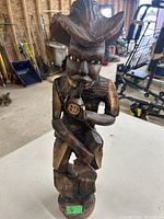 Front view of a wood carving figurine sitting on a table in a workshop area, showing detailed carving of the face, hat, and hands holding a pipe.