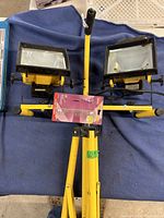 Two yellow portable halogen work lights on a collapsible tripod stand, one bulb installed, the other missing bulb, and an extra sealed bulb resting on the stand.