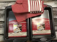 Set of 2 Wilton baking pans, red oven mitt and red striped tea towel shown from top angle on wooden surface.