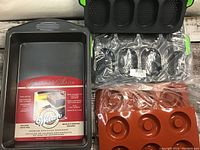 Photo showing a Wilton branded rectangular metal cake pan with packaging insert, alongside three silicone baking molds of different shapes and colors arranged on a wooden surface.