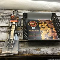 BBQ grill basket with product label showing BBQ food images placed on wooden surface next to stainless steel grilling utensils in packaging.