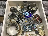 Top-down photo showing various pieces of costume jewelry including bracelets, earrings, brooches, and pendants all arranged loosely inside a tray.