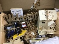 Photo of the craft box contents showing a wooden 'welcome' sign, small decorative house, framed signs, paintbrushes, glue device and sticks, red paint jar, and twine, packed in cardboard box with shredded filler.