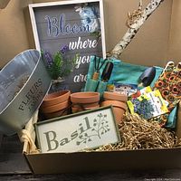 Overview of garden box items showing pots, tools, seed packets, gloves, metal tote, and decorative signs arranged inside a cardboard box.