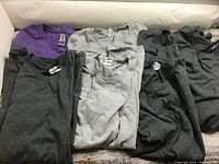 Six folded Gildan Softstyle T-shirts in dark gray, light gray, and purple colors, neatly arranged in a box.